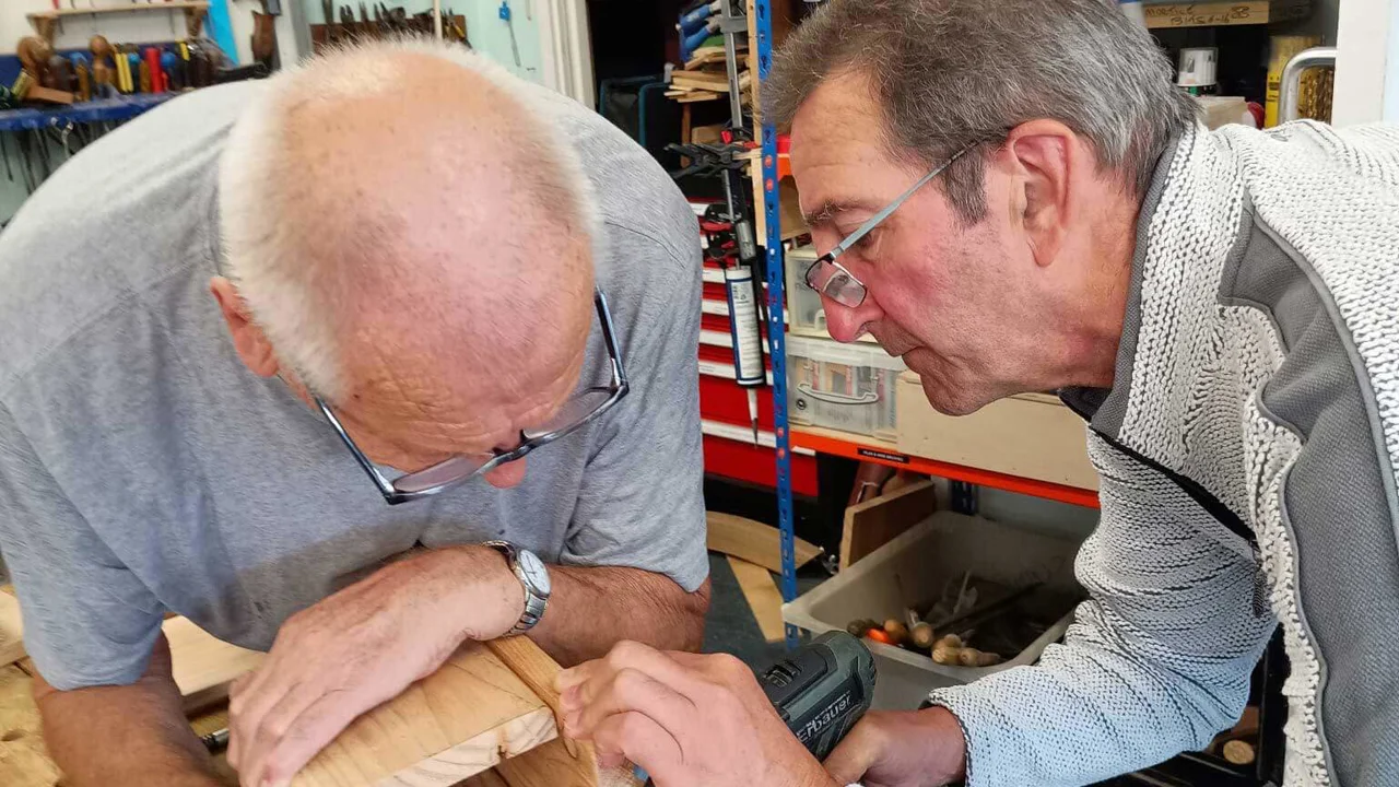 Two mean leaning over a wood construction, one is drilling and the other holding the wood both concentrating at Corsham Men's shed
