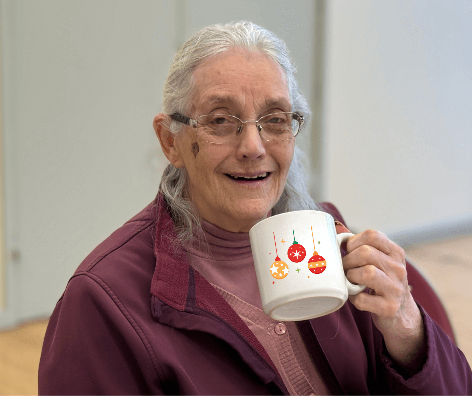 Ann in a purple jacket, with a festive mug in hand, smiling at the camera at Kennet Muddy Boots group