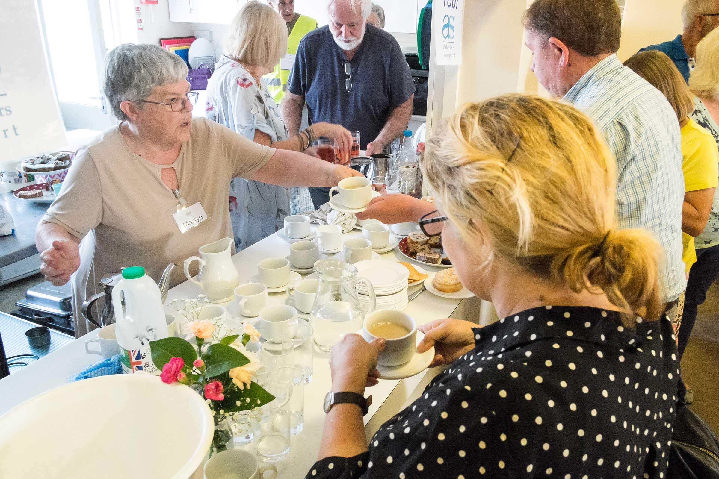 A older lady is helping a gentleman who is holding a cup of tea, from behind a table which has empty cups and saucers with another lady in a spotty top holding a cup of tea.