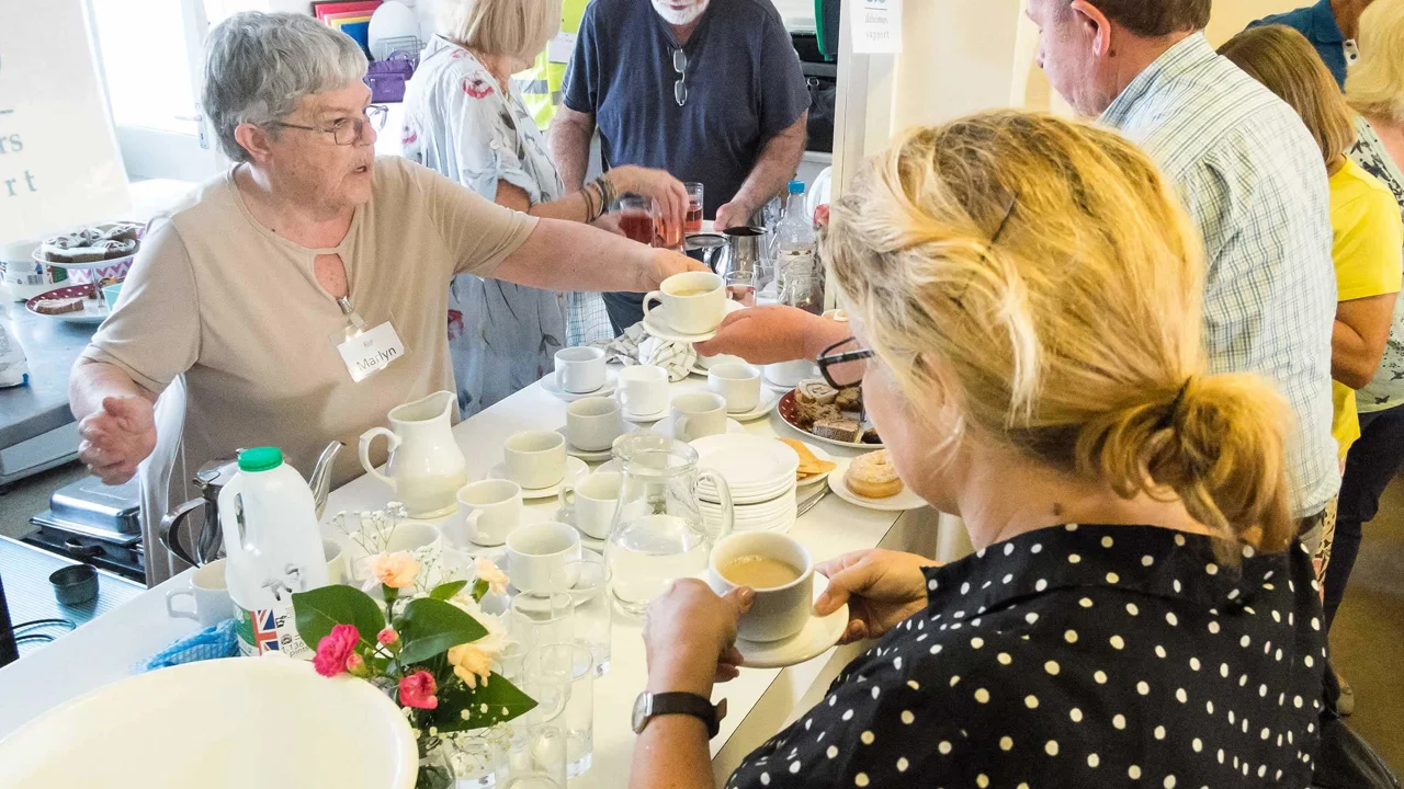 A older lady is helping a gentleman who is holding a cup of tea, from behind a table which has empty cups and saucers with another lady in a spotty top holding a cup of tea.