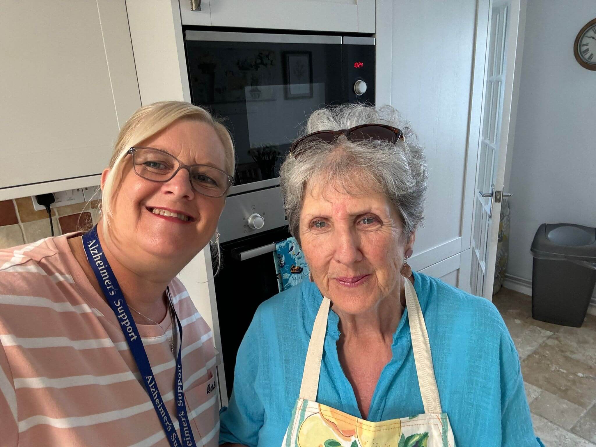 Two ladies stand together in a kitchen, one is wearing a pink stripy top and glasses, the other a blue top and an apron with glasses on her head. They are both smiling at the camera