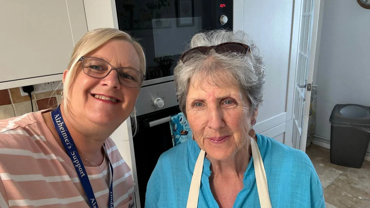 Two ladies stand together in a kitchen, one is wearing a pink stripy top and glasses, the other a blue top and an apron with glasses on her head. They are both smiling at the camera