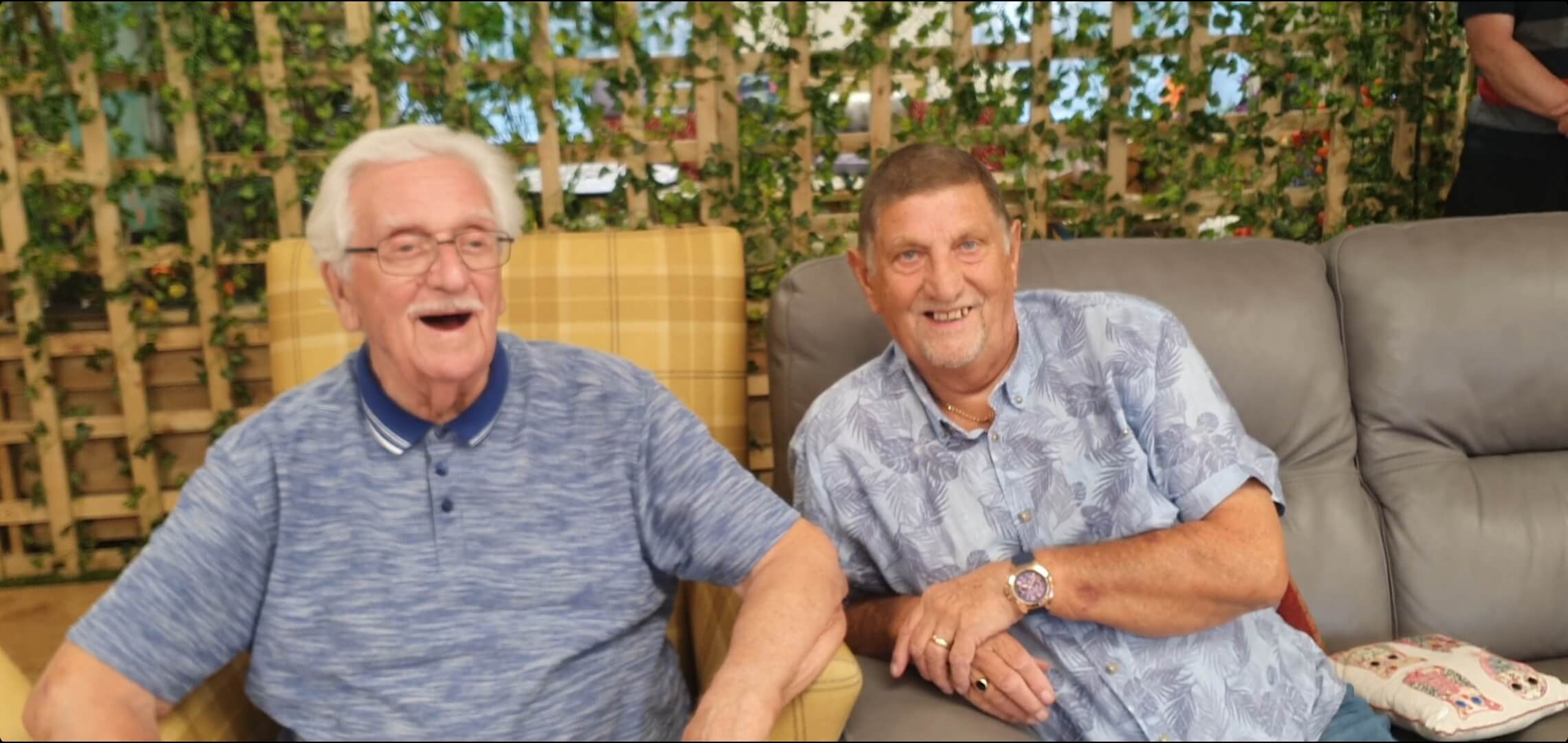 Two gentlemen sitting next to each other in arm chairs at Old Silk Works club, one has a grey t shirt and his mouth open laughing, the other in a blue shirt is leaning towards the other smiling 