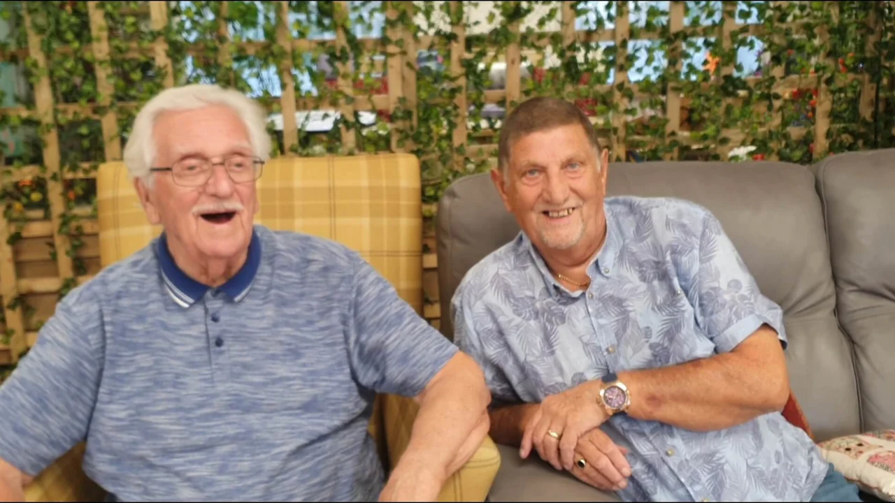 Two gentlemen sitting next to each other in arm chairs at Old Silk Works club, one has a grey t shirt and his mouth open laughing, the other in a blue shirt is leaning towards the other smiling