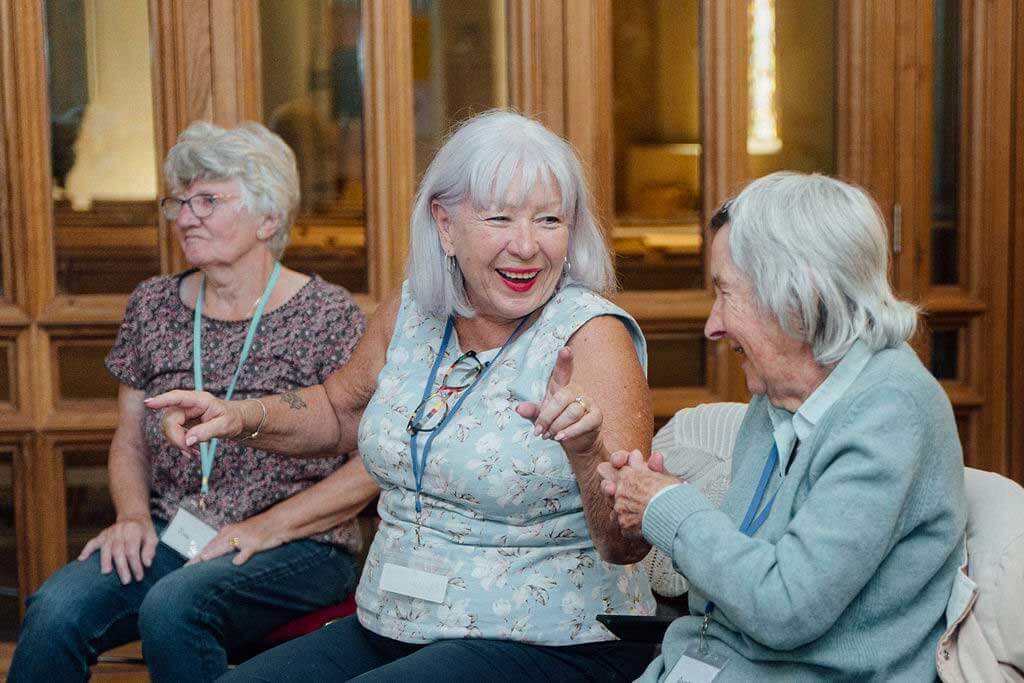 Two ladies with grey hair in front of picture sharing a funny moment and laughing with a lady behind watching something across the room