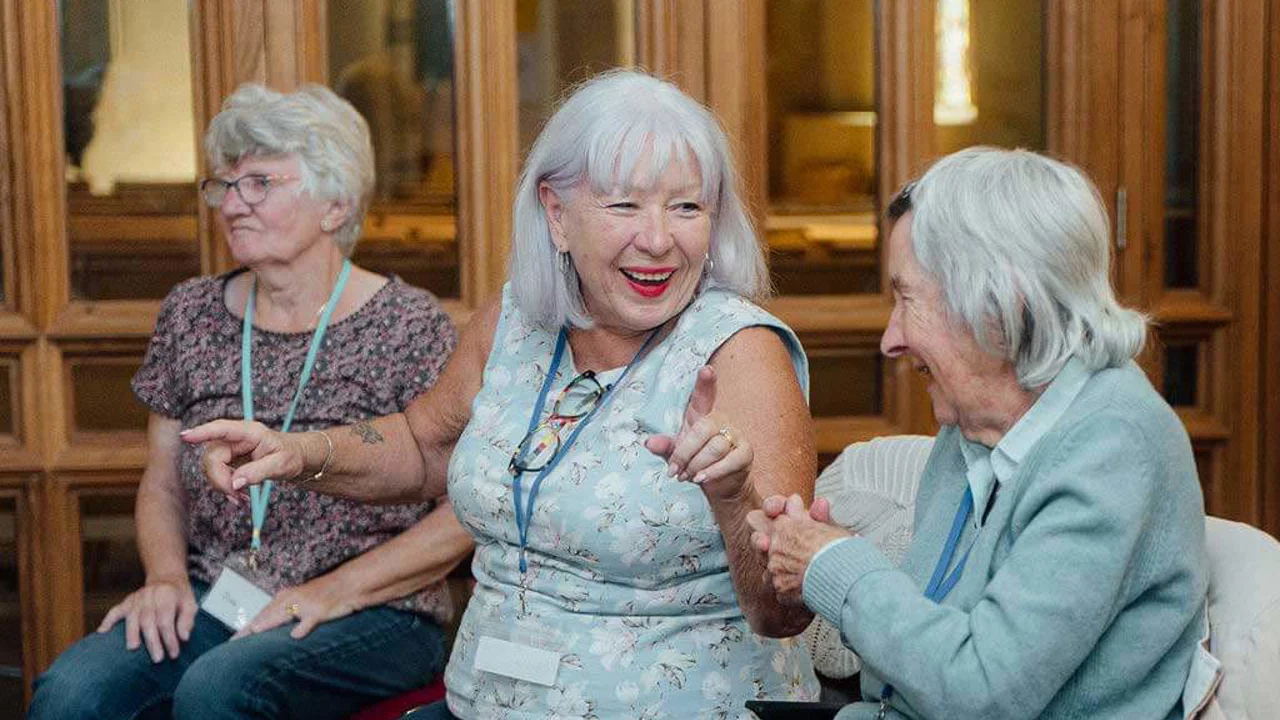 Two ladies with grey hair in front of picture sharing a funny moment and laughing with a lady behind watching something across the room