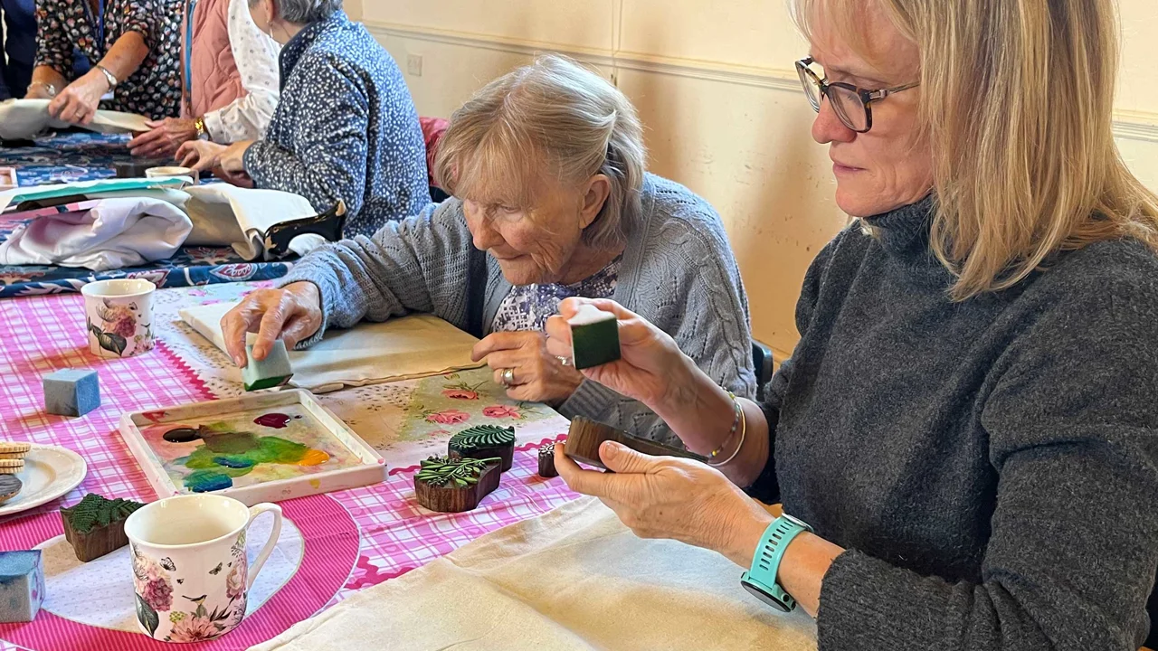 A lady in a grey jumper is in the foreground adding paint to a stencil stamp. The lady next to her in pale grey leans forward to reach the paint. There are three other ladies chatting together and a table full of paint, cups and fabric.