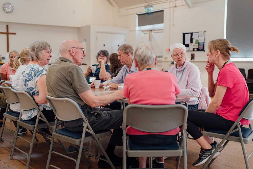 A group of older men and women sitting round a table with cups of tea, chatting to a younger woman in a t shirt and trainers 