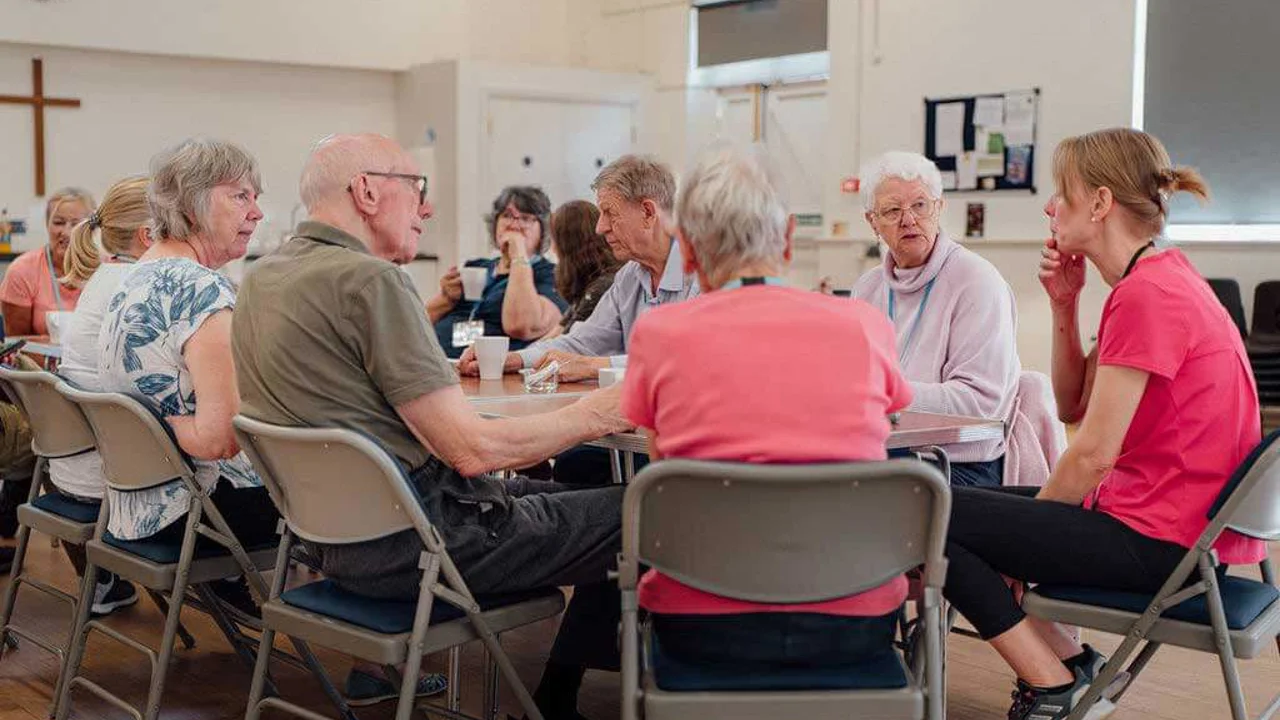A group of older men and women sitting round a table with cups of tea, chatting to a younger woman in a t shirt and trainers