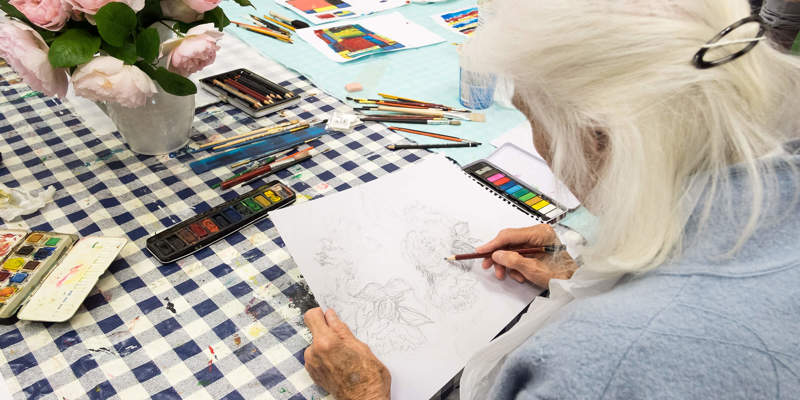 Woman with long white head drawing a still life of roses in pencil. Taken from over her shoulder so we see the back of her head and sketch emerging