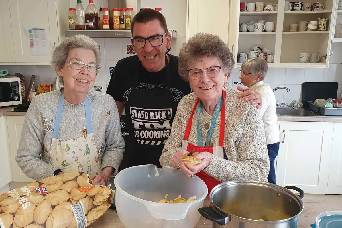 Two ladies and a man standing close together smiling whilst potato peeling with large saucepan and potatoes in the foreground.