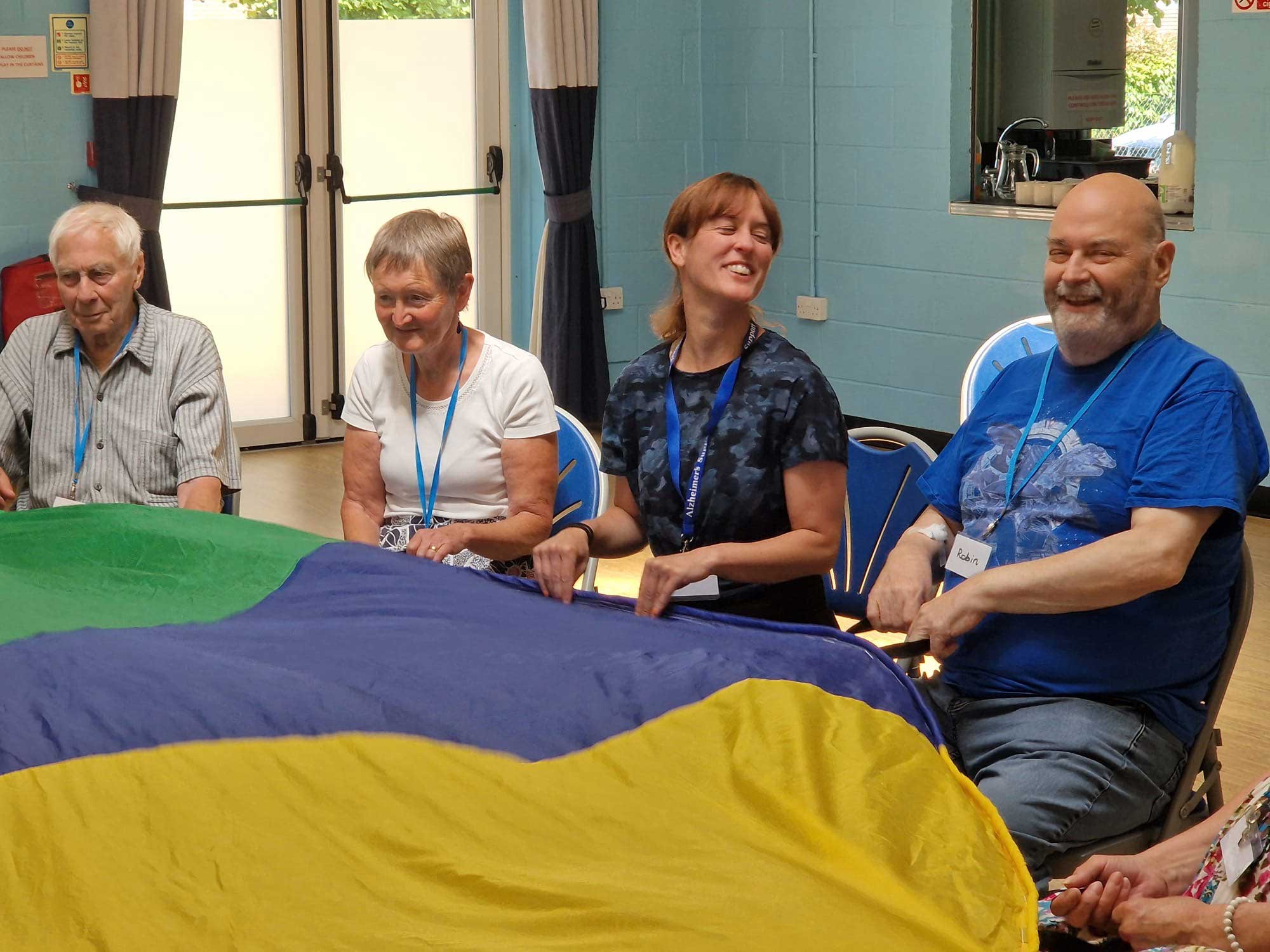 Four people holding a blue, yellow and green parachute in front of them, all smiling at Melksham Movement for the Mind 