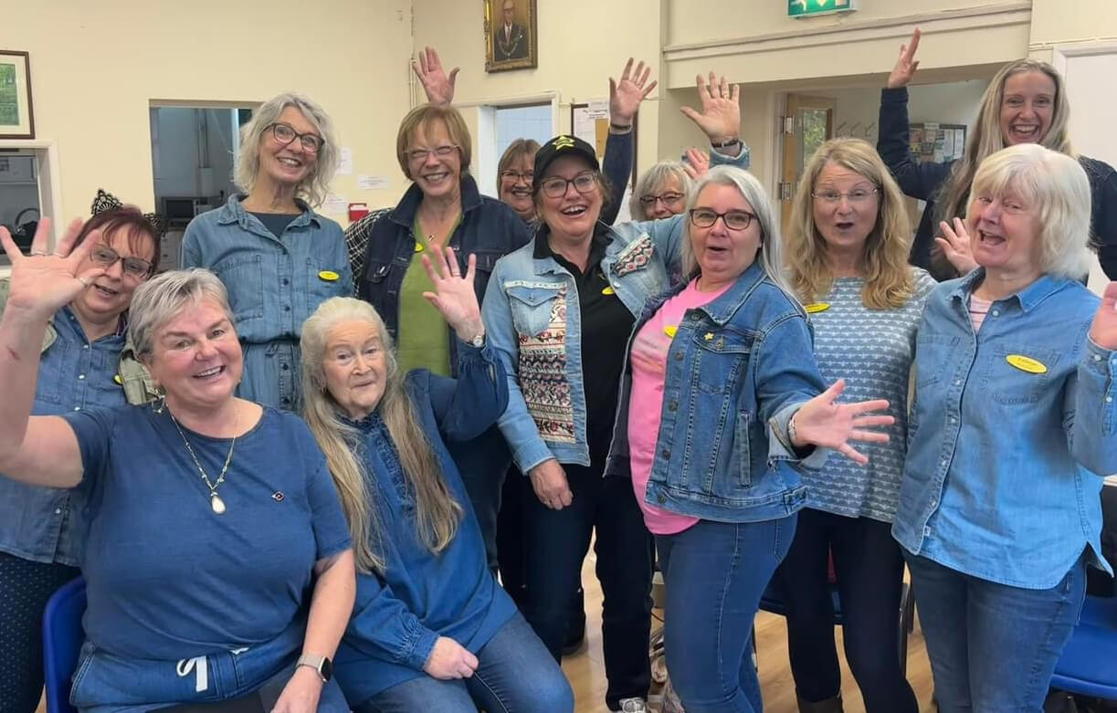 A group of ladies all wearing Denim for Dementia blue, standing, sitting, waving and smiling at the camera 