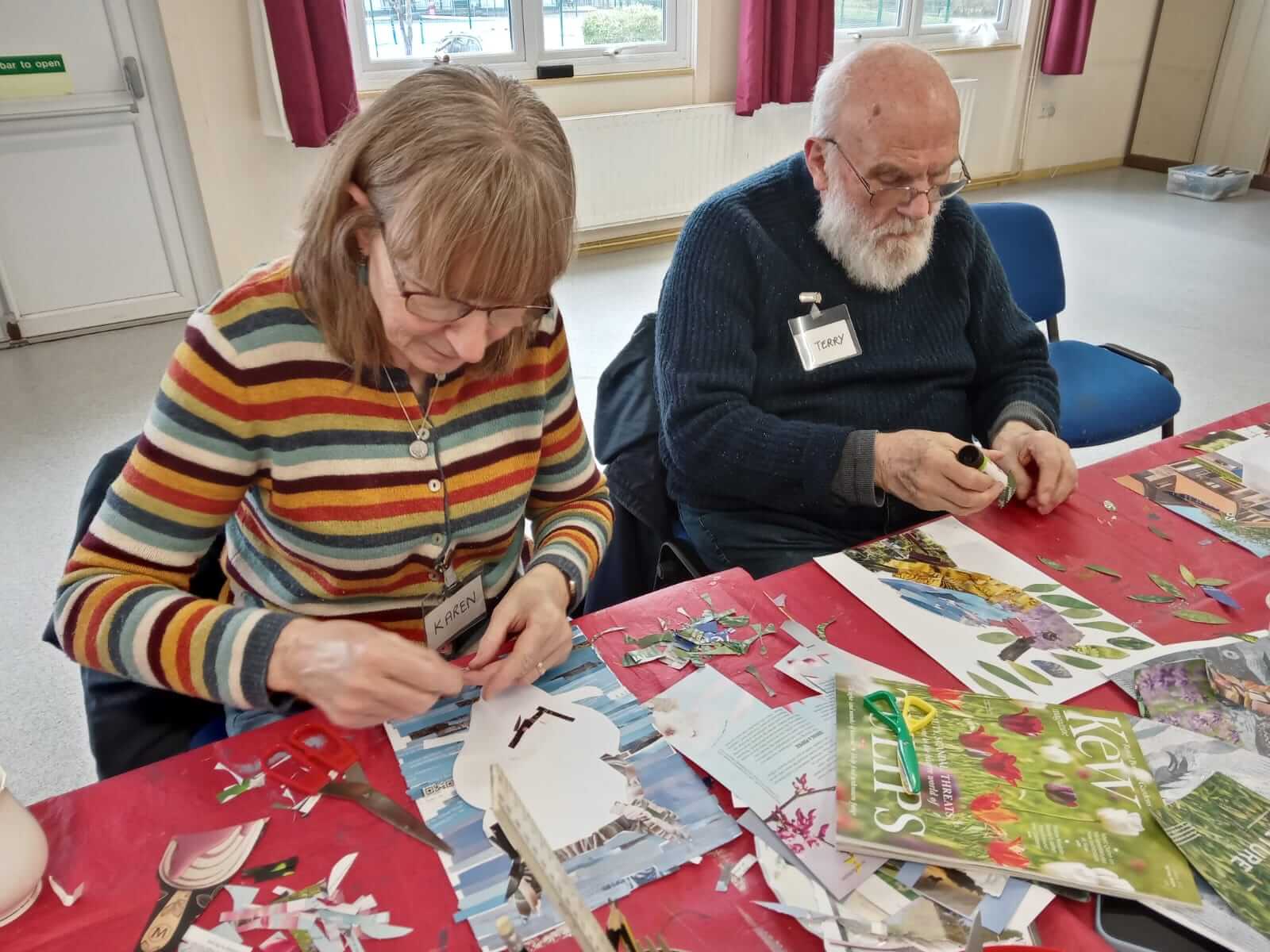 A lady wearing a stripy cardigan is sitting at a table next to a man in a blue jumper.  They are both sticking paper to a picture making a collage.  Taken at Warminster Art Group 