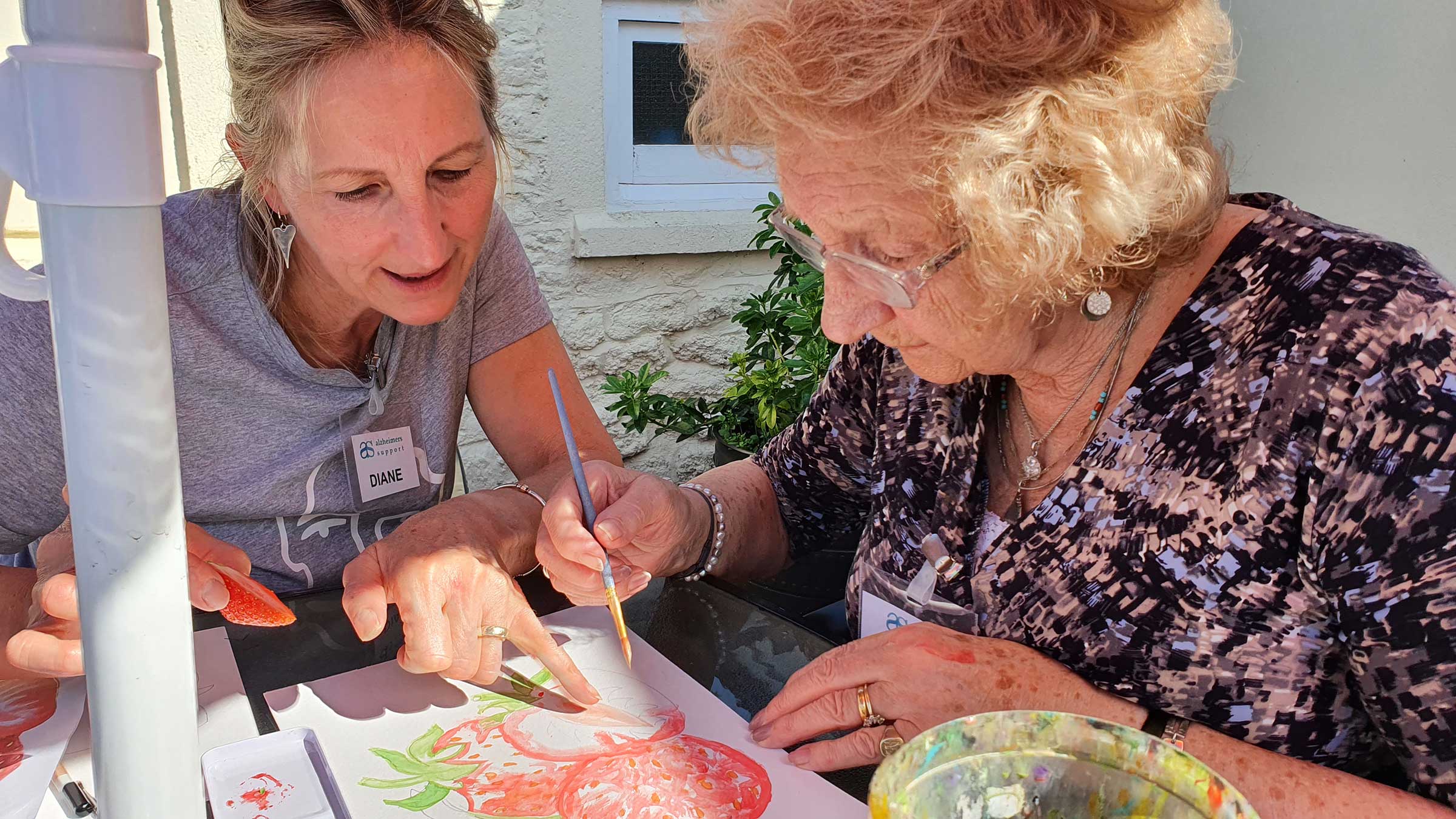 An older lady is painting a picture of red strawberries, whilst a lady wearing grey points to her picture next to her .  They are sitting outside on a sunny day at Lyneham Art group 