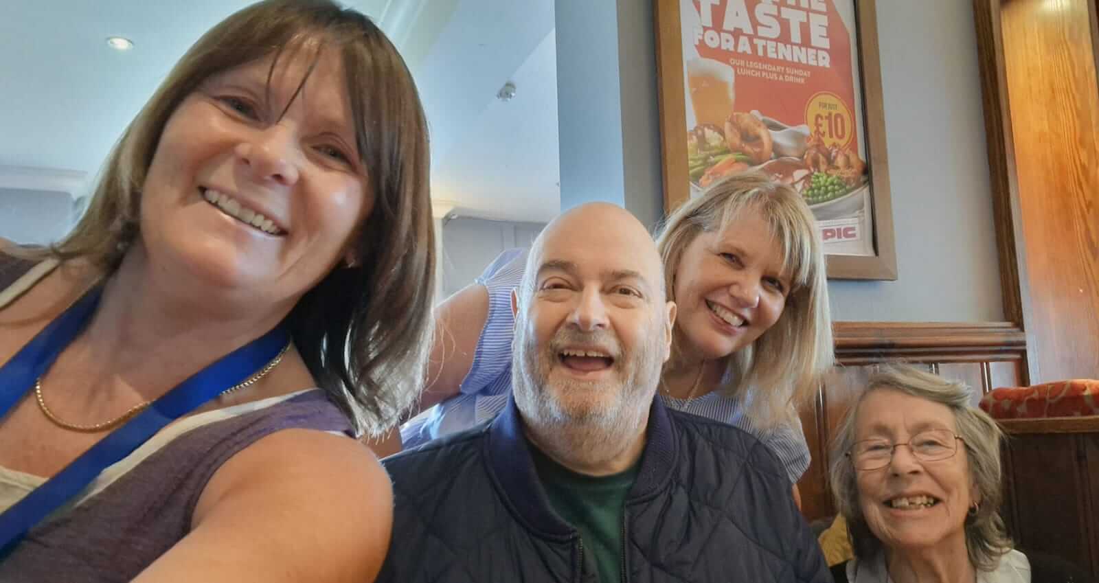 One lady with brown hair and a vest top is smiling next to a man with a bald head and beard, next to a lady with glasses and grey hair, behind them a lady with blond hair leans in for the picture. They are seated in a restaurant and all smiling