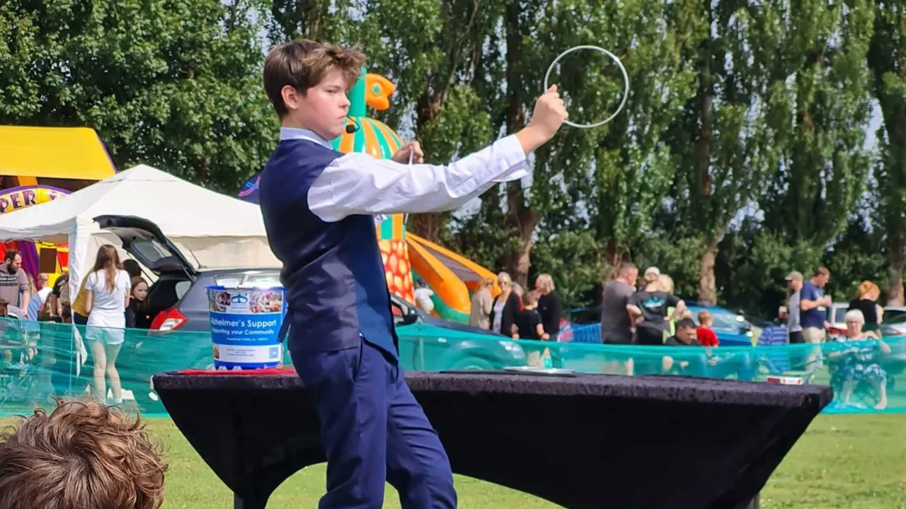 A young magician wearing a waistcoat standing in a park setting holding silver hoops and doing a magic trick to people watching in the foreground
