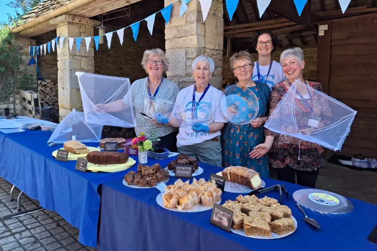 Group of lovely smiling ladies on a sunny day in front of a stone barn with blue bunting, standing behind a cake table with eight different cakes and blue table cloths.