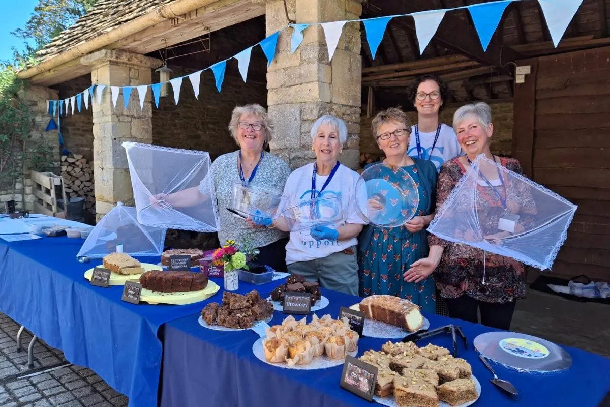 Group of lovely smiling ladies on a sunny day in front of a stone barn with blue bunting, standing behind a cake table with eight different cakes and blue table cloths.