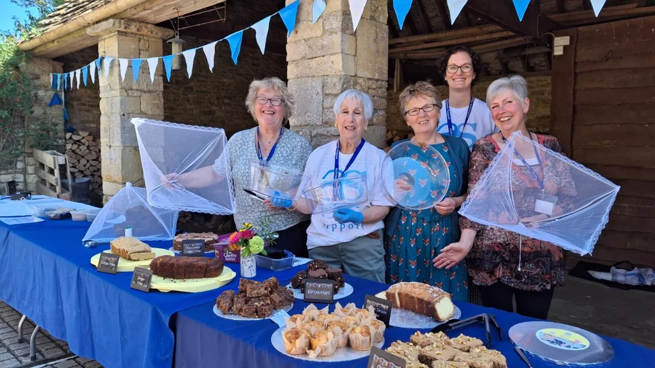 Group of lovely smiling ladies on a sunny day in front of a stone barn with blue bunting, standing behind a cake table with eight different cakes and blue table cloths.