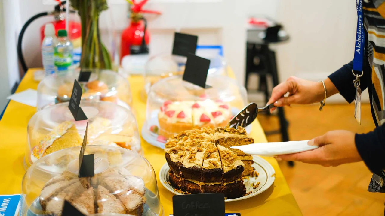 This shows a table top with a display of cakes, there is a lady holding a cake slice and helping herself to a slice of cake of Coffee and Walnut cake