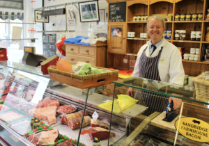 The picture shows a butchers shop, Chris Robbins is standing smiling behind the glass counter with meats and a pay machine, behind him there are wooden shelves with jars, there is a wooden chest to the right before a window with a view out to the street.