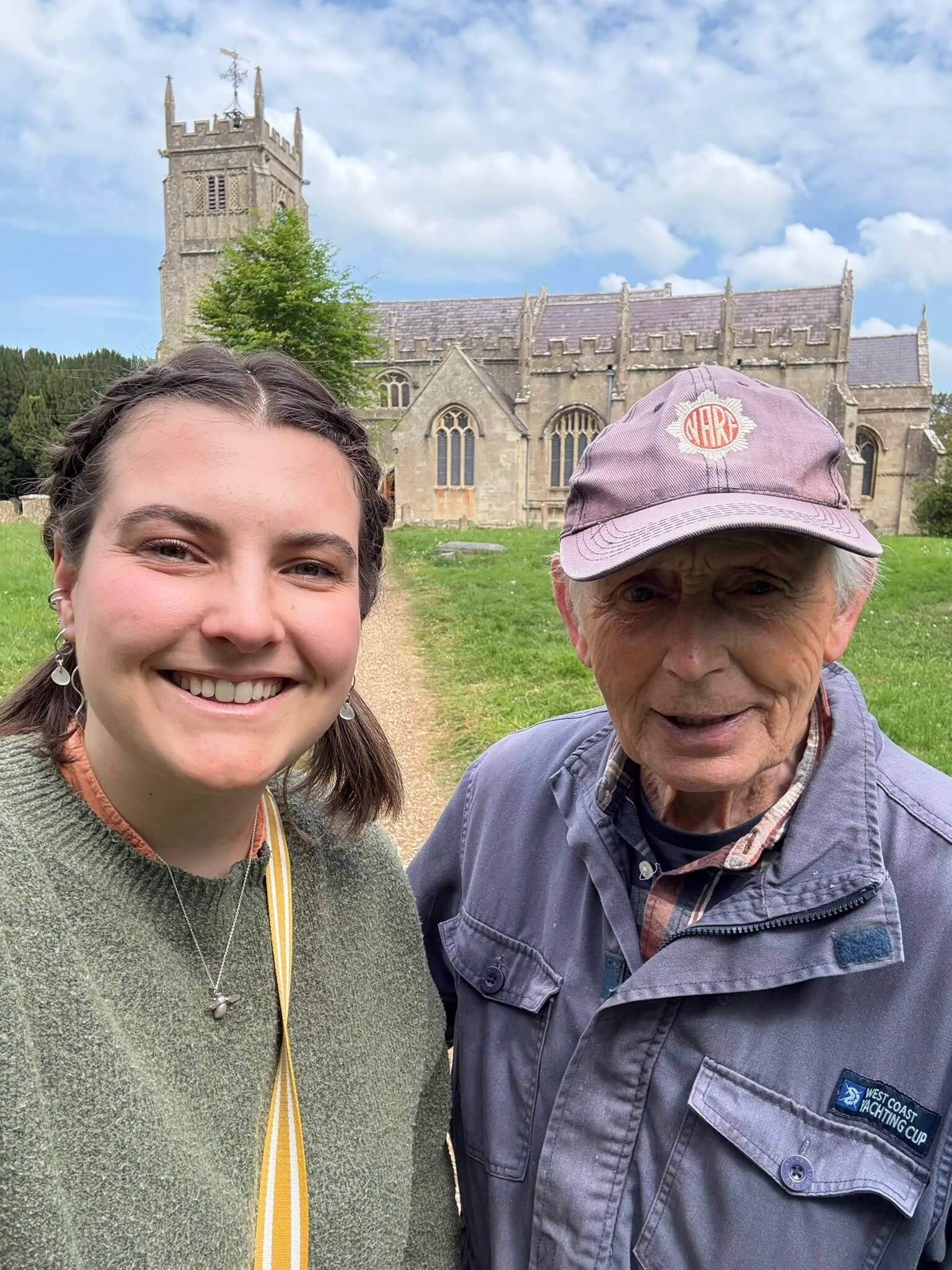 On the right is an older gentleman in a baseball cap and a blue jacket.  On the left is a younger lady in a grey jumper and dark hair.  They are standing in front of a beautiful church