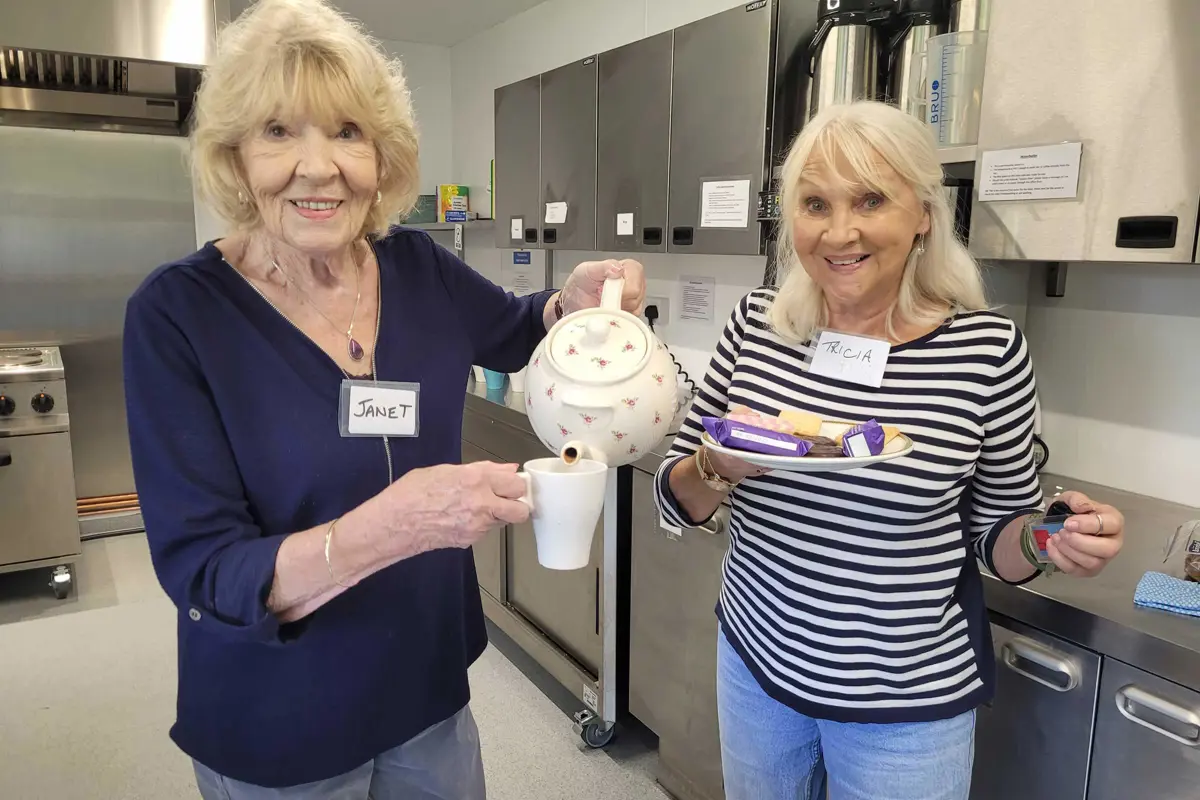 Two smiling volunteers are standing in a kitchen, one in a blue top is pouring tea from a pot to a cup and the other in a stripy top holds a plate of biscuits