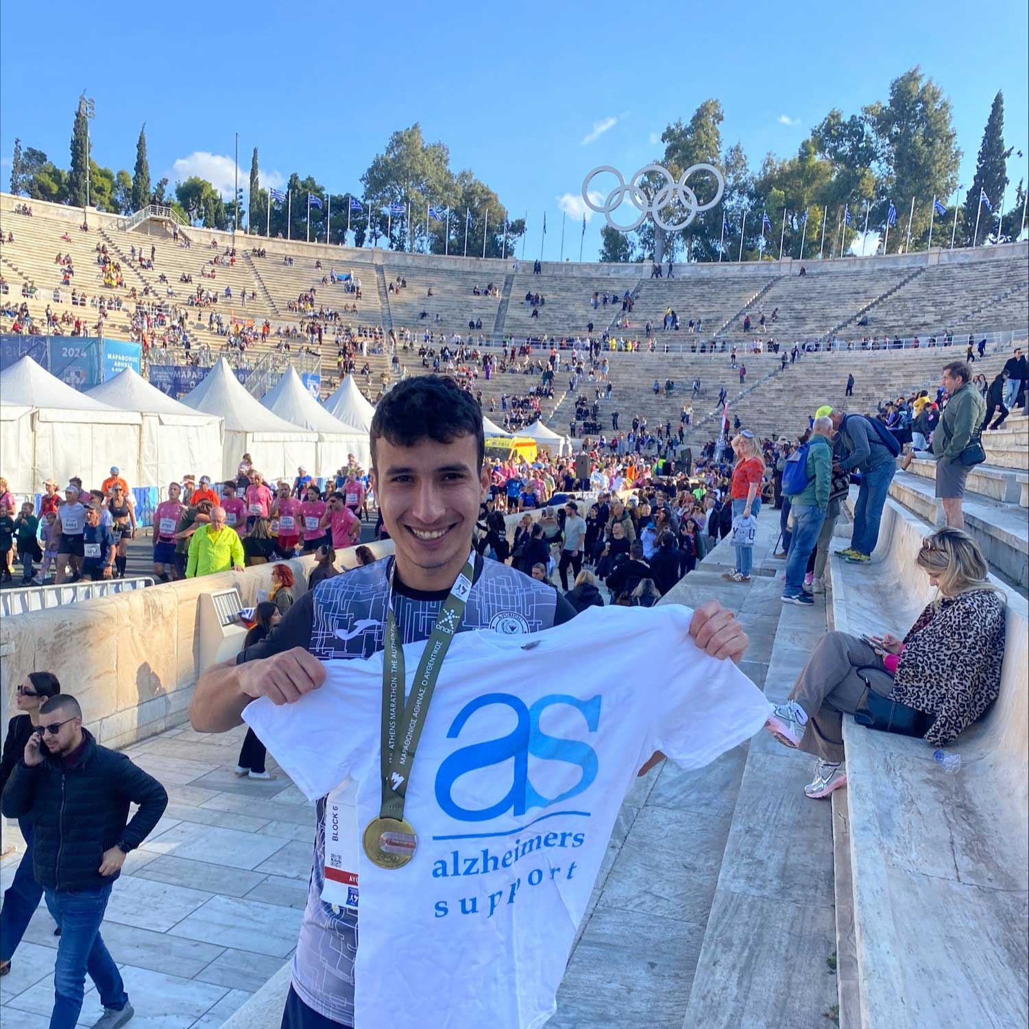 A young man with dark hair, wearing a gold medal and holding a white Alzheimer's T shirt is standing a huge amphitheatre with tents and people walking and sitting.  There are hundreds of stone steps and at the top are the five Olympic rings and flags
