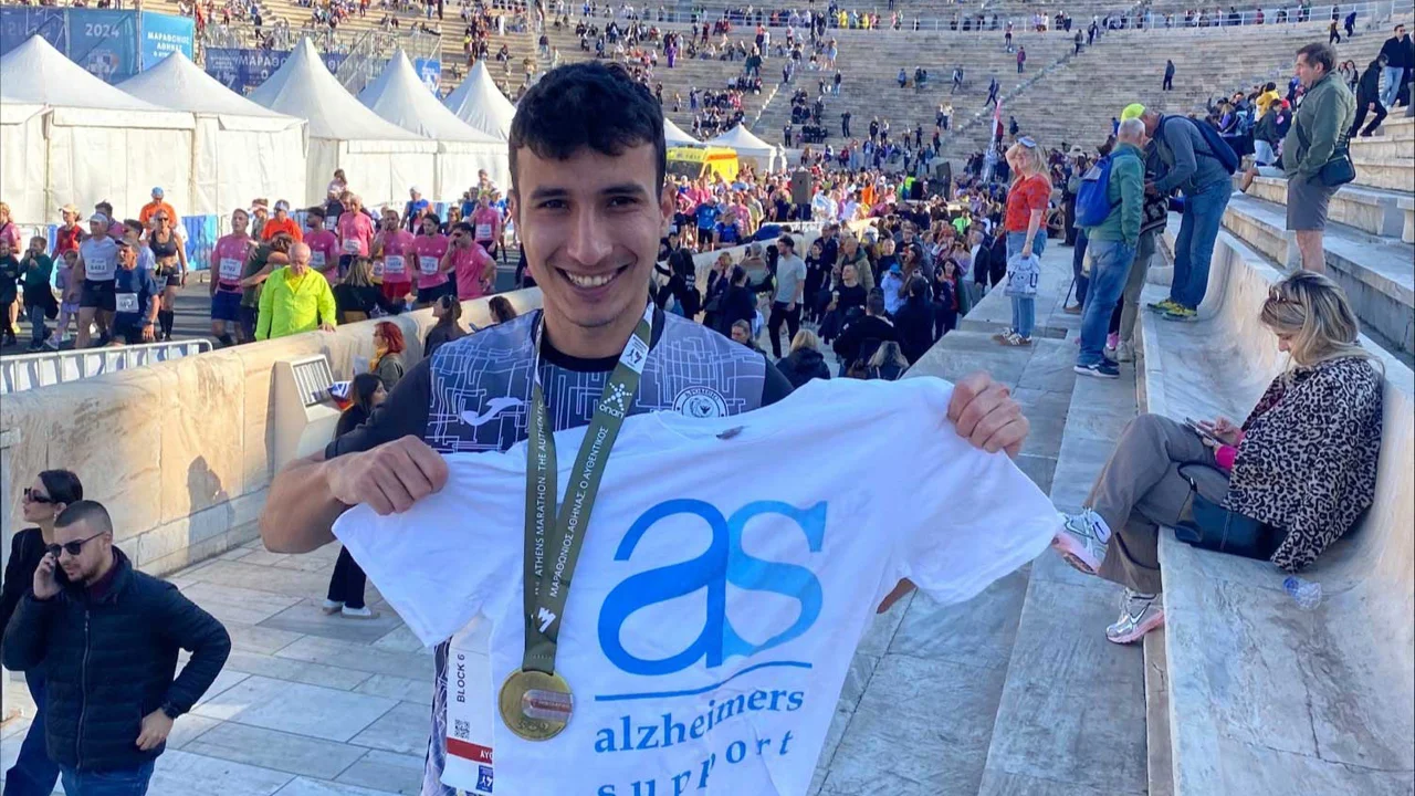 A young man with dark hair, wearing a gold medal and holding a white Alzheimer's T shirt is standing a huge amphitheatre with tents and people walking and sitting. There are hundreds of stone steps and at the top are the five Olympic rings and flags