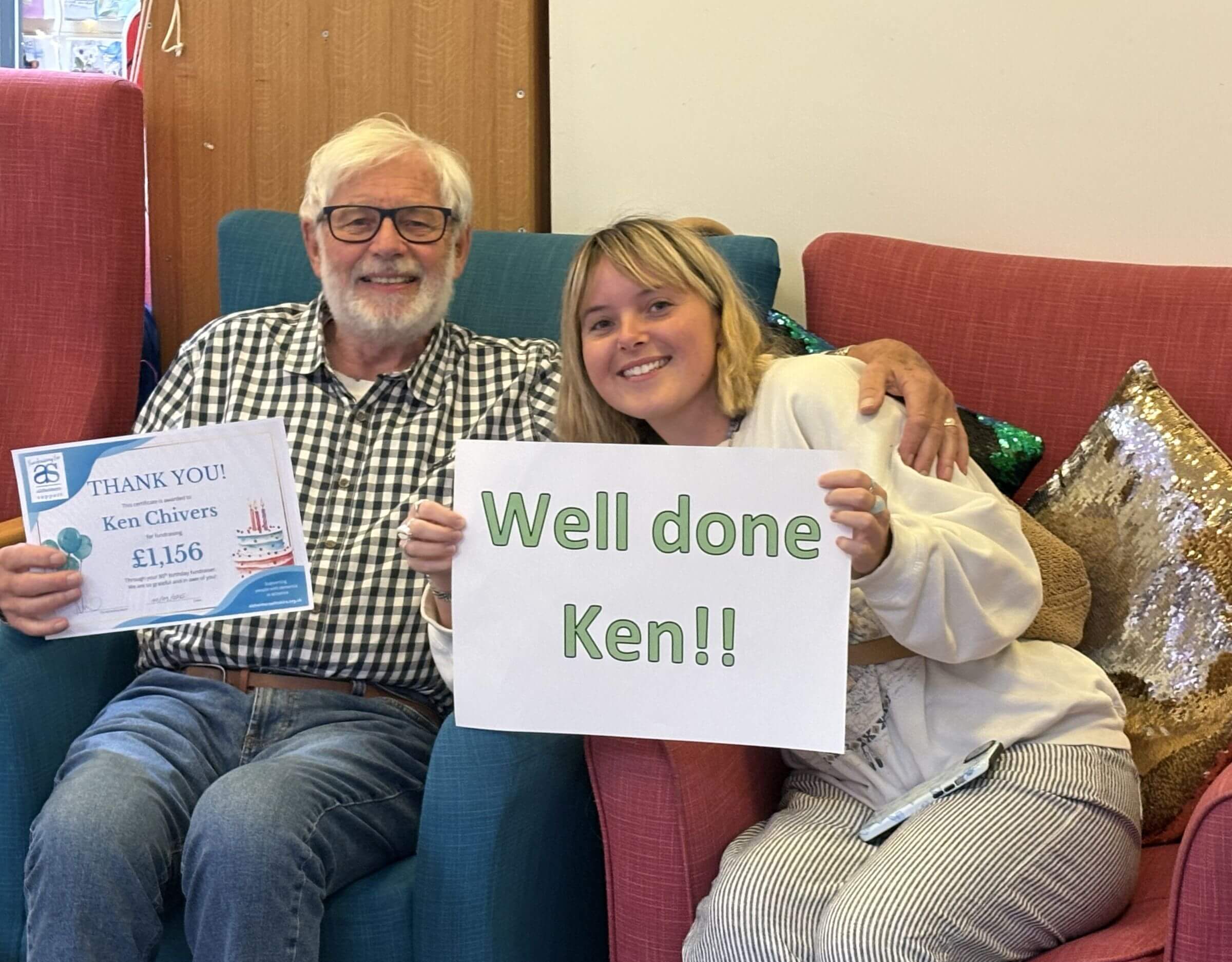 Ken in a checked shirt and his daughter sitting in armchairs close together. He is holding a thank you certificate and she a Thank You Ken sign.