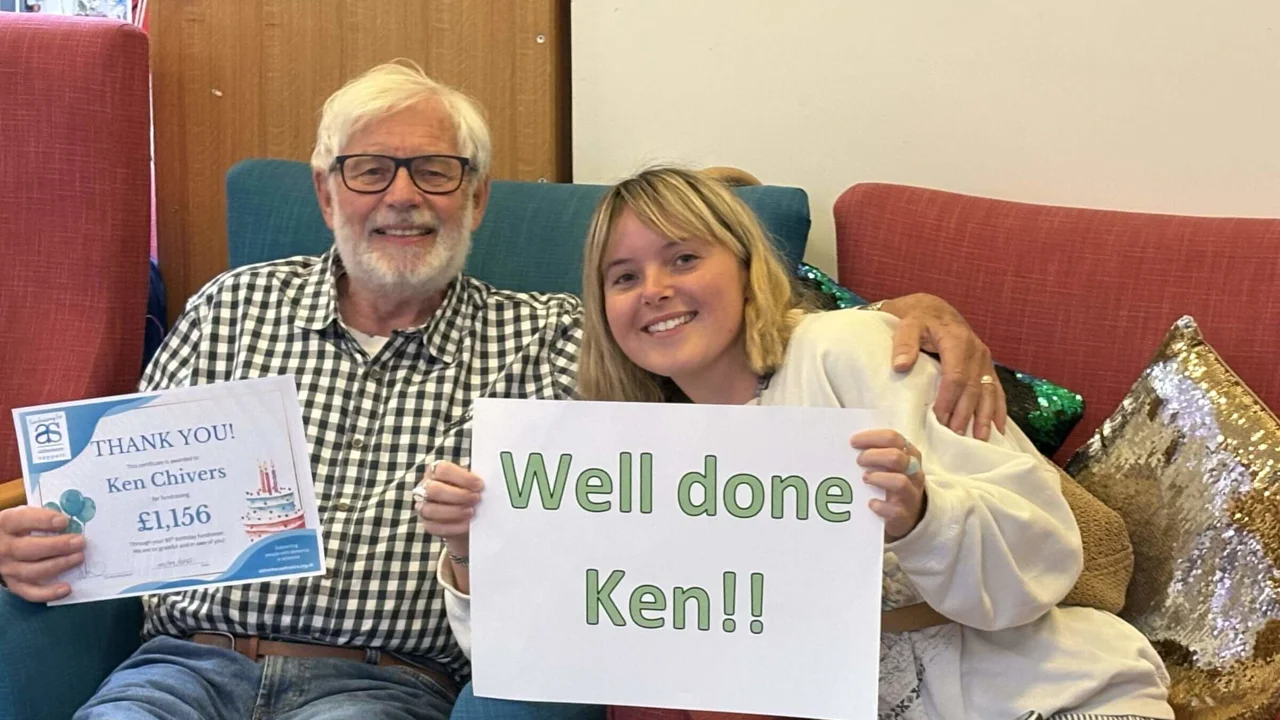 Ken in a checked shirt and his daughter sitting in armchairs close together. He is holding a thank you certificate and she a Thank You Ken sign.