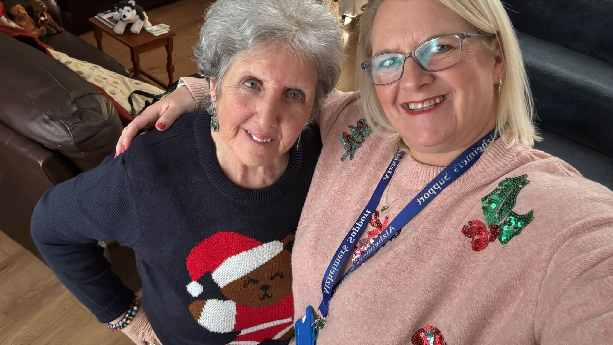 Two ladies are wearing Christmas jumpers - one is blue with a teddy dressed as Santa on, the other is pink with holly berries in green and red.  She is standing with her arm round the other lady and smiling