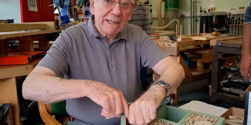 Man wearing a grey t shirt, seated at a work bench making an insect hotel by putting tubes into a shape of wood at the Corsham Men's Shed