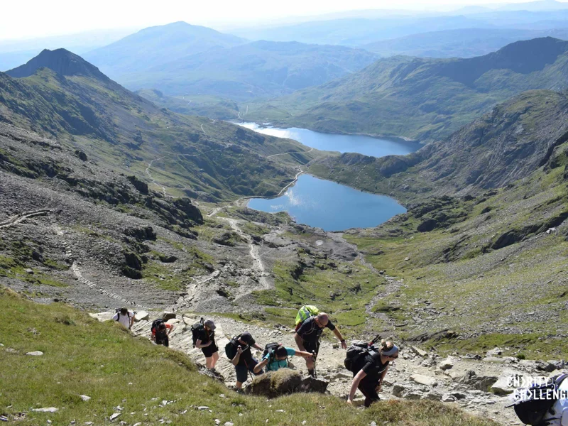 This shows a huge rugged, mountainous scenery with people in t shirts and walking gear climbing up a steep stony path in the foreground with two lakes visible in the distance, the sun is shining. This shows a huge rugged, mountainous scenery with people in t shirts and walking gear climbing up a steep stony path in the foreground with two lakes visible in the distance, the sun is shining.