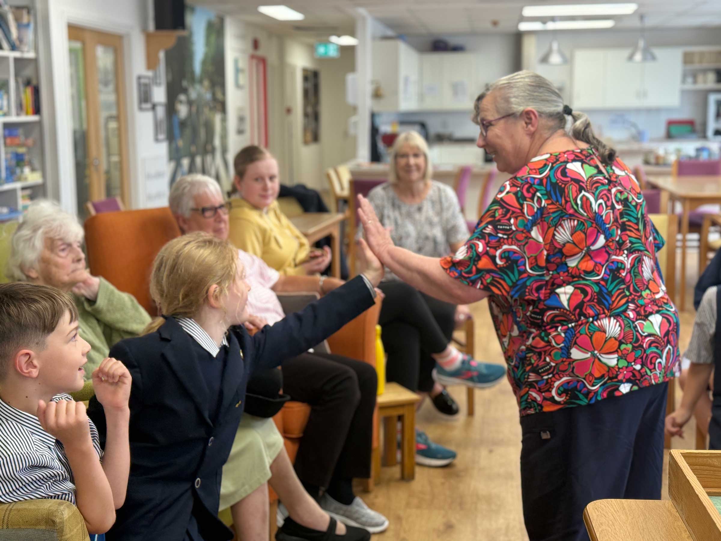An older lady is standing facing a line of ladies sat in arm chairs with two younger children in school uniform sat with them,  The lady is high fiving the younger girl and both are smiling 