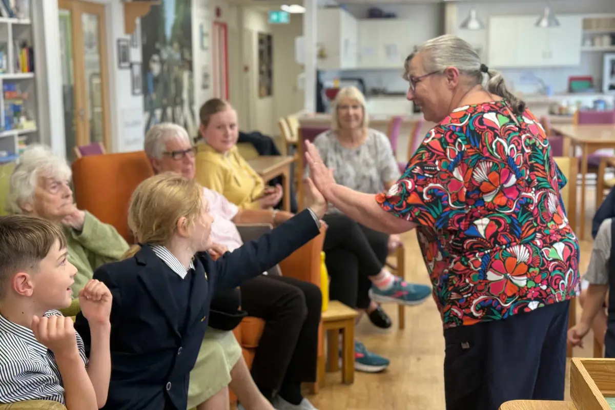 An older lady is standing facing a line of ladies sat in arm chairs with two younger children in school uniform sat with them,  The lady is high fiving the younger girl and both are smiling 