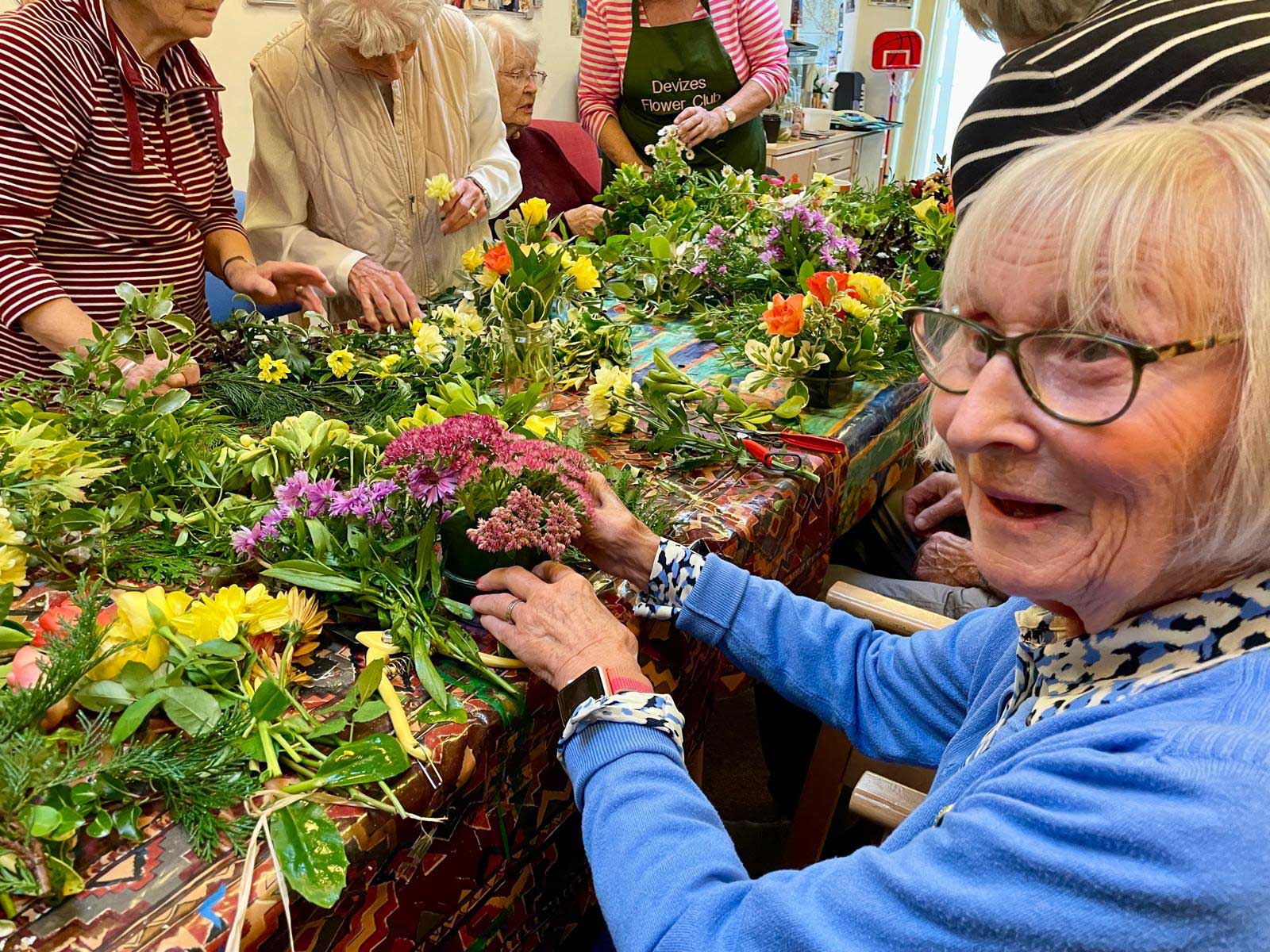 Older lady wearing blue smiling whilst flower arranging pink and purple flowers in Sidmouth Street 
