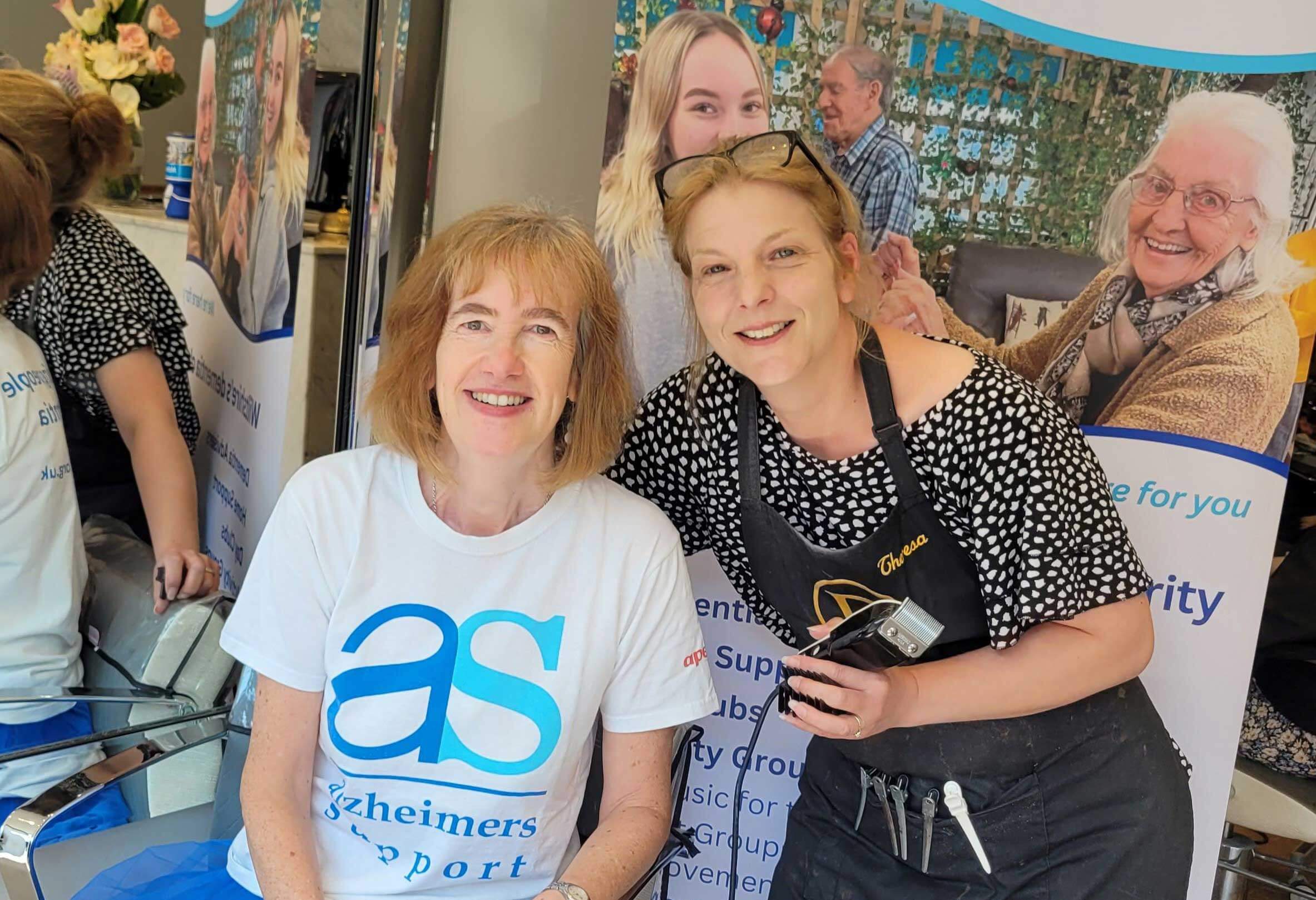 Sarah T sitting in a hair dressers wearing a AS T shirt with another lady in a spotty top holding clippers about to give her a buzz cut to raise charity funds