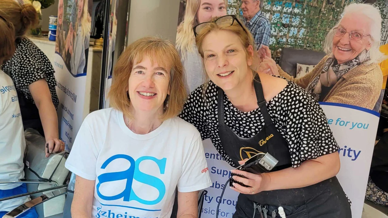 Sarah T sitting in a hair dressers wearing a AS T shirt with another lady in a spotty top holding clippers about to give her a buzz cut to raise charity funds