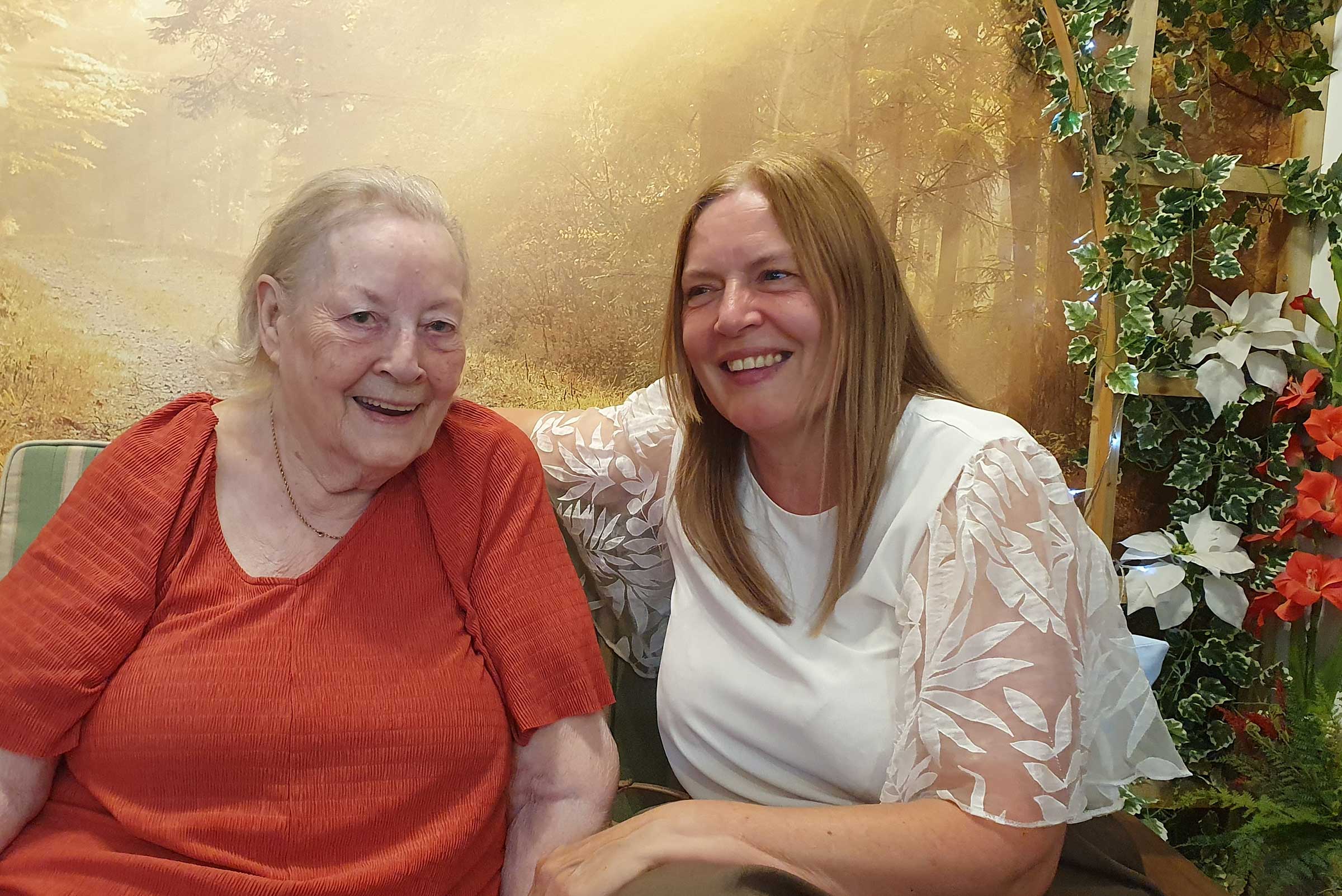 Jean, in a red top, sitting with her daughter Julie in white, on a bench in an indoor garden area with foliage around. Both smiling.