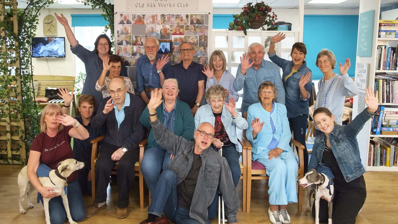 Group of club members staff and volunteers, some seated, some standing, wearing their demin outfits and waving for the camera