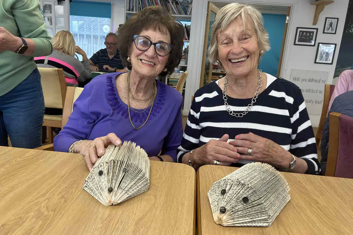 One lady in a blue top and one lady in a stripy top sitting at a table making paper hedgehogs, both smiling at the camera in Old Silk Works club 