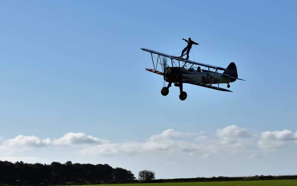 A picture of blue sky with a bi-plane in silhouette with someone doing a Wing Walk and a person piloting the plane with a field and trees at the bottom