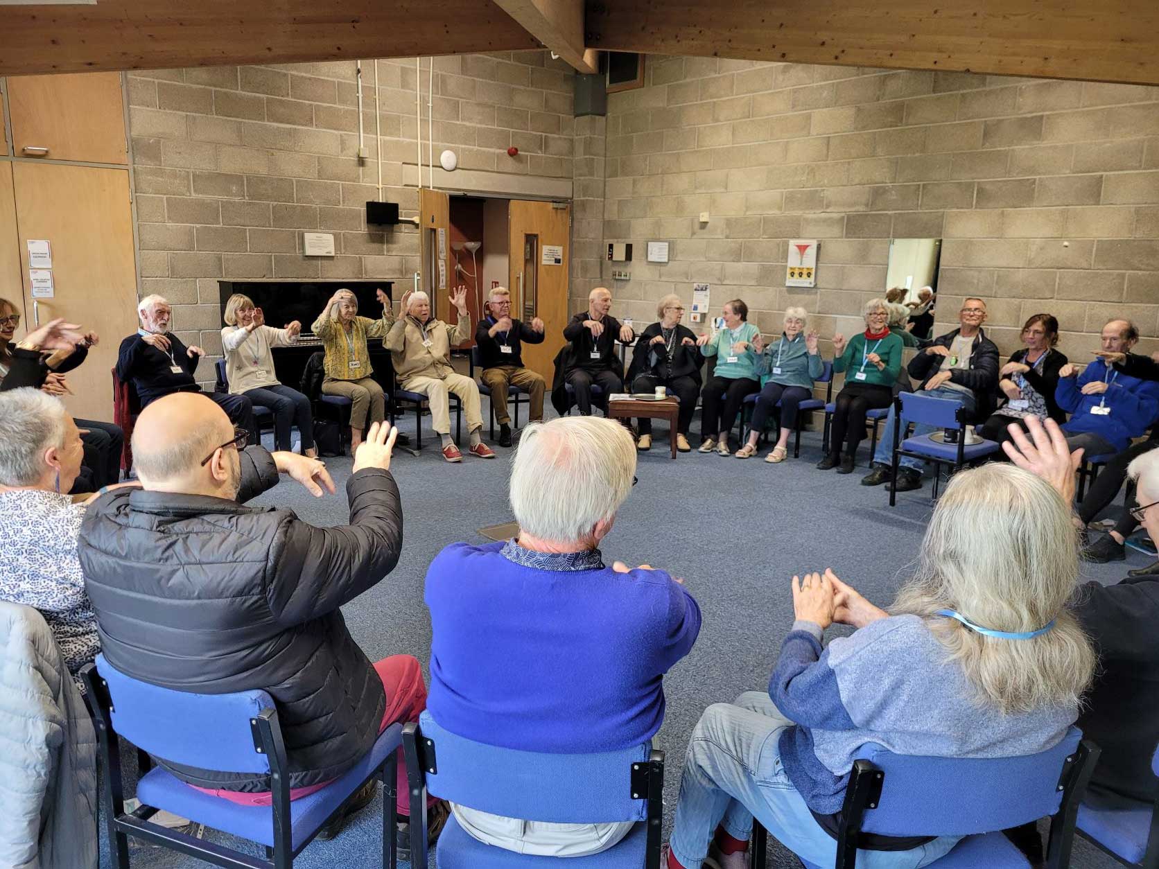 A large group of men and women all seated in a circle in a Hall.  They are all raising their arms in the same movement at Bradford Music for the Mind 