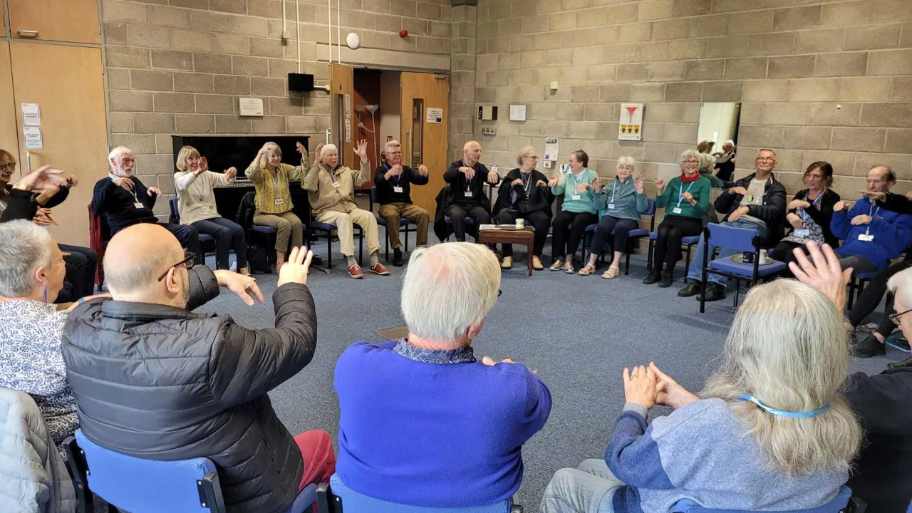 A large group of men and women all seated in a circle in a Hall. They are all raising their arms in the same movement at Bradford Music for the Mind