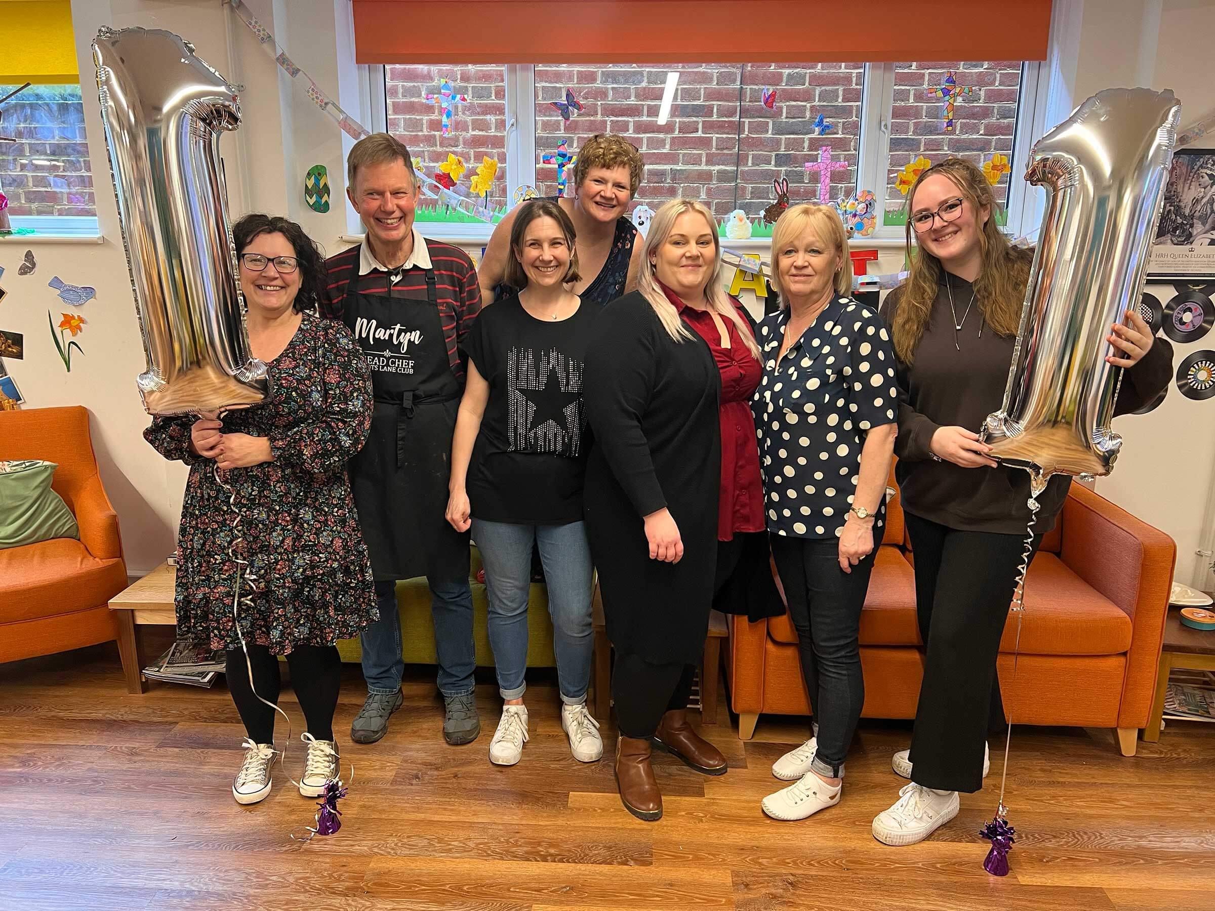 Group of smiling staff and volunteers holding large silver balloons in the shape of a number 1, inside Scots Lane club