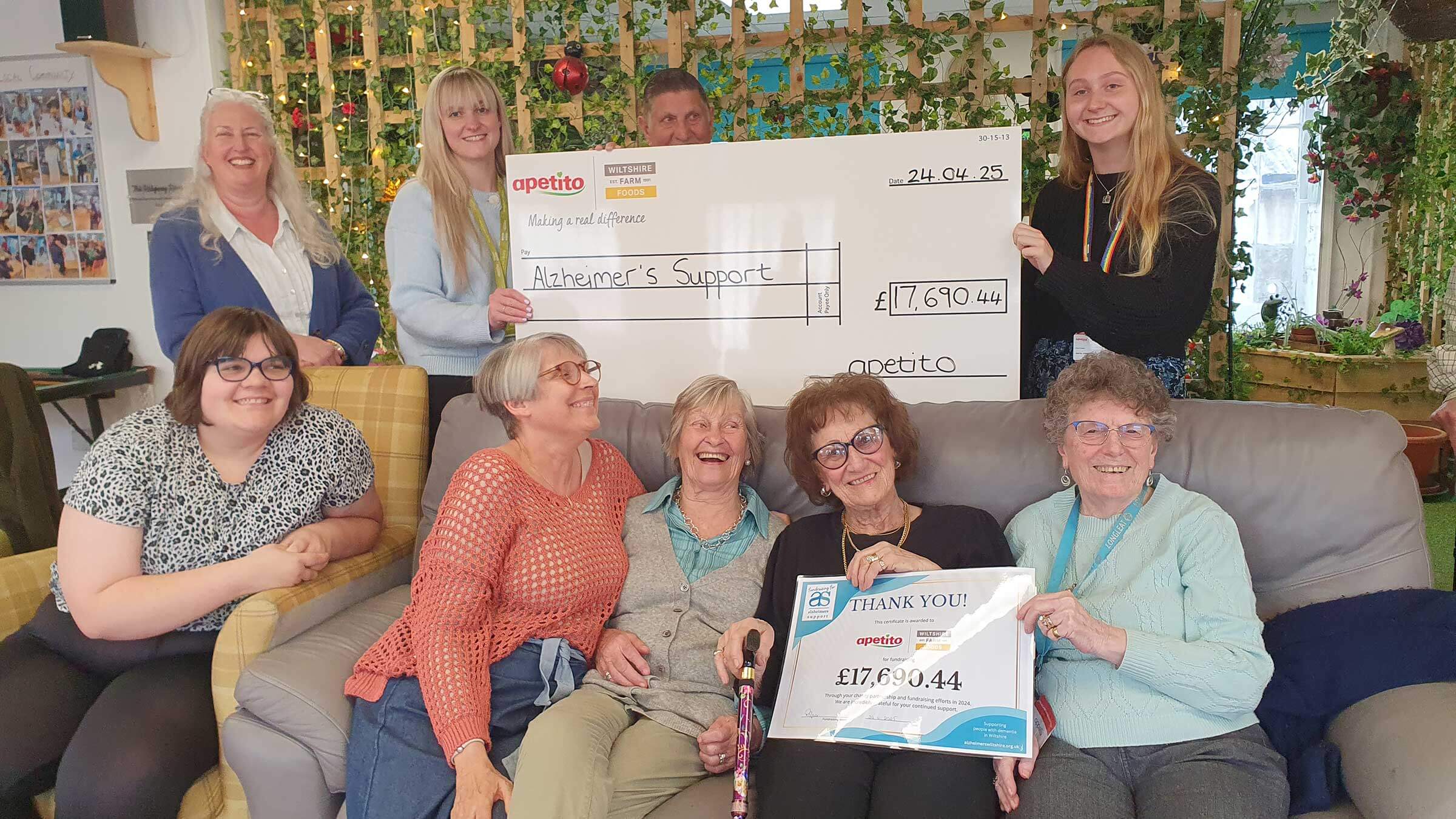 A group of day club members with staff and apetito representatives. Some are sitting together on a sofa holding a thankyou certificate, with others behind holding a giant cheque