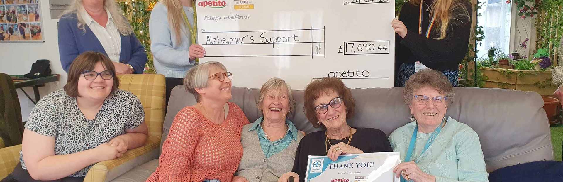 A group of day club members with staff and apetito representatives. Some are sitting together on a sofa holding a thankyou certificate, with others behind holding a giant cheque A group of day club members with staff and apetito representatives. Some are sitting together on a sofa holding a thankyou certificate, with others behind holding a giant cheque