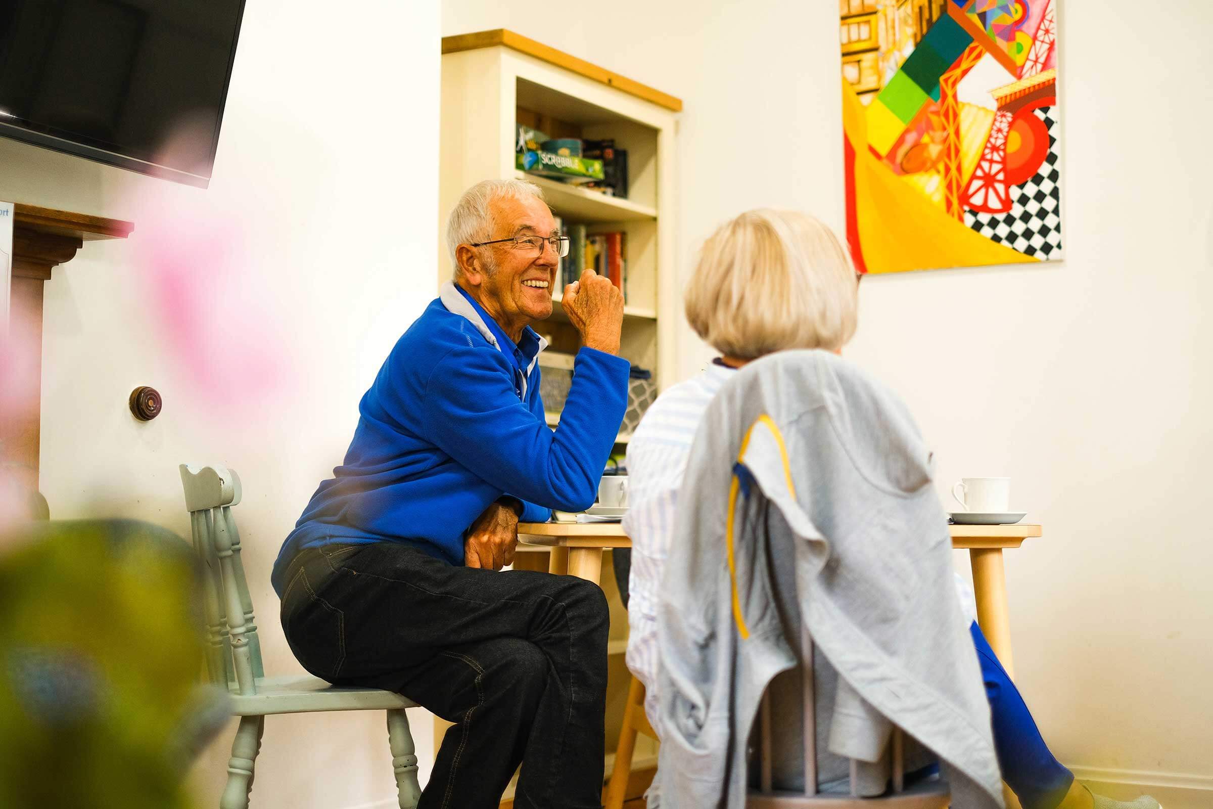 A man sitting at a table in a cafe with a book shelf and a picture of modern art with a lady whose back is to us.  He has a blue jumper and is laughing and chatting.